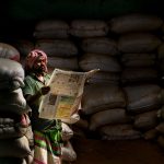 A man in traditional attire reads a newspaper in a dimly lit storeroom with stacked sacks. — QBet Casino im internationalen V