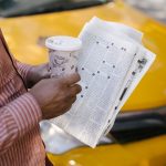 From above side view of crop anonymous ethnic male cab driver with coffee to go and newspaper talking to coworker on street —